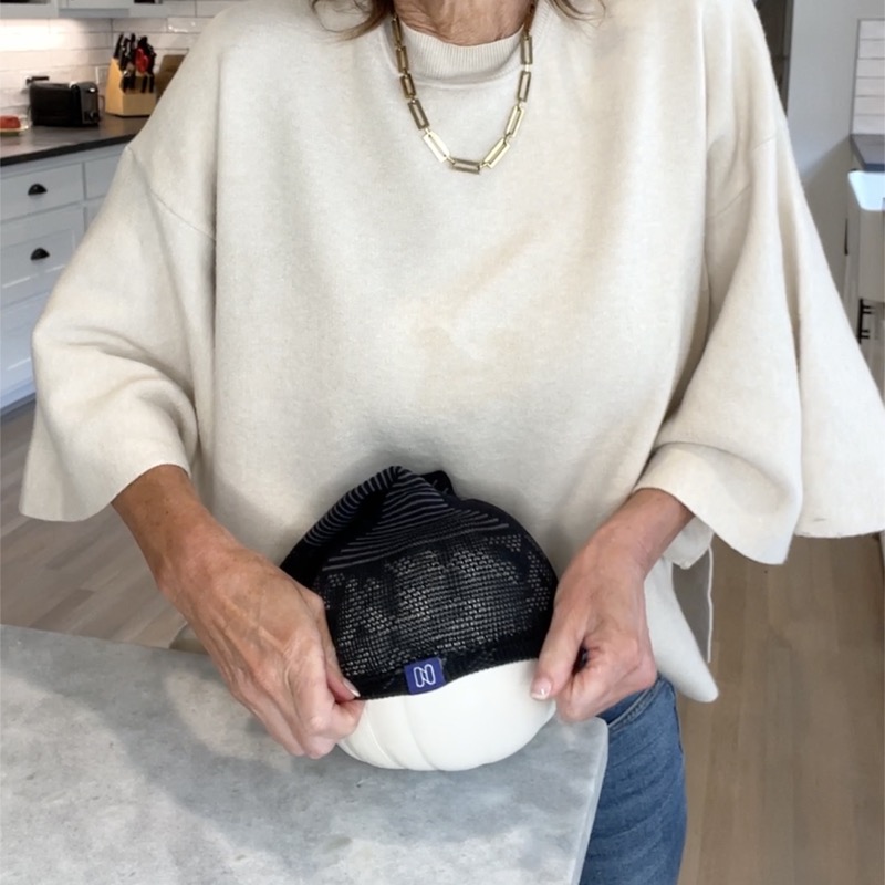 A woman in a cream sweater, photographed from the chin down in a home kitchen, holding a black NanoSocks compression sock stretched over a white decorative pumpkin to show how far the knit opens up.